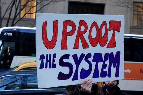 A person holds up a sign that reads "Uproot the system"