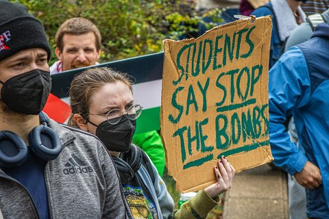 A person wearing a mask and glasses holds a sign reading "Students say stop the bombs"