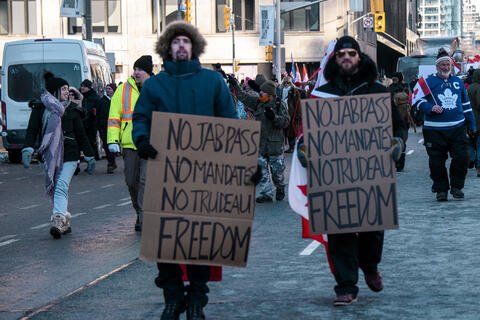 Two people in winter clothes holding signs that say "No jab pass, no mandates, no Trudeau, Freedom"