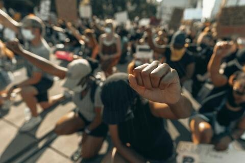 Focus on a raised fist, background of people kneeling in protest.