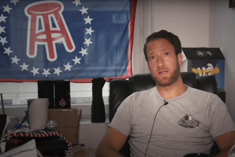 A man in a grey T-shirt sitting in a chair with a flag behind him with the Barstool logo