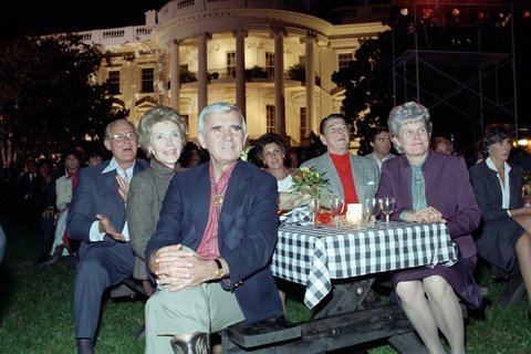 Six people sitting around a gingham table cloth on a picnic table with the white house in the background