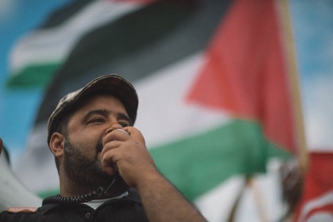 man speaking into bullhorn in front of palestinian flag
