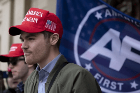 a man in a green jacket and a Make America Great Again hat with an America First flag behind him.
