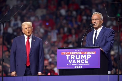 rfk jr at podium with trump vance sign, trump looks on