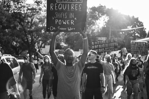a person holding a sign that reads "dismantling a racist system requires shifts in power - Dr. Sherry Watt"