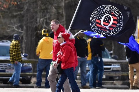 People gathering for QAnon rally with two people holding flag in front of trucks
